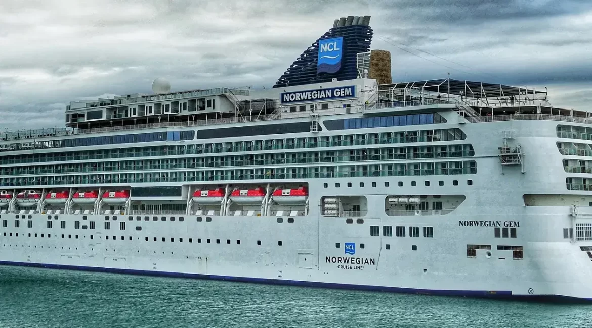 A large cruise ship, the Norwegian Gem, docked in a harbor on a cloudy day.