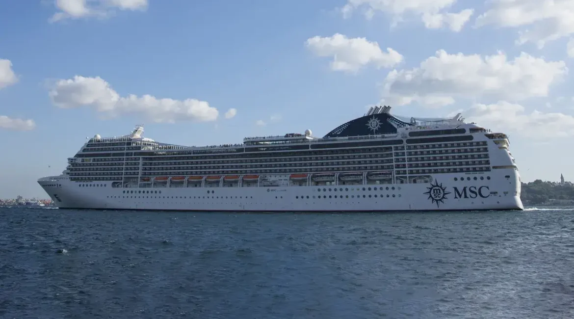 Large MSC cruise ship sailing under a partly cloudy blue sky, with the coastline visible in the background.